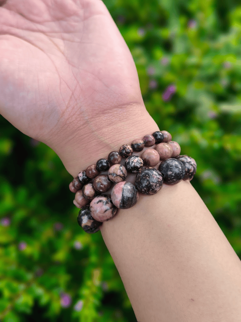 Pink and black Rhodonite round-bead bracelets in several bead sizes, worn together on a wrist outdoors.