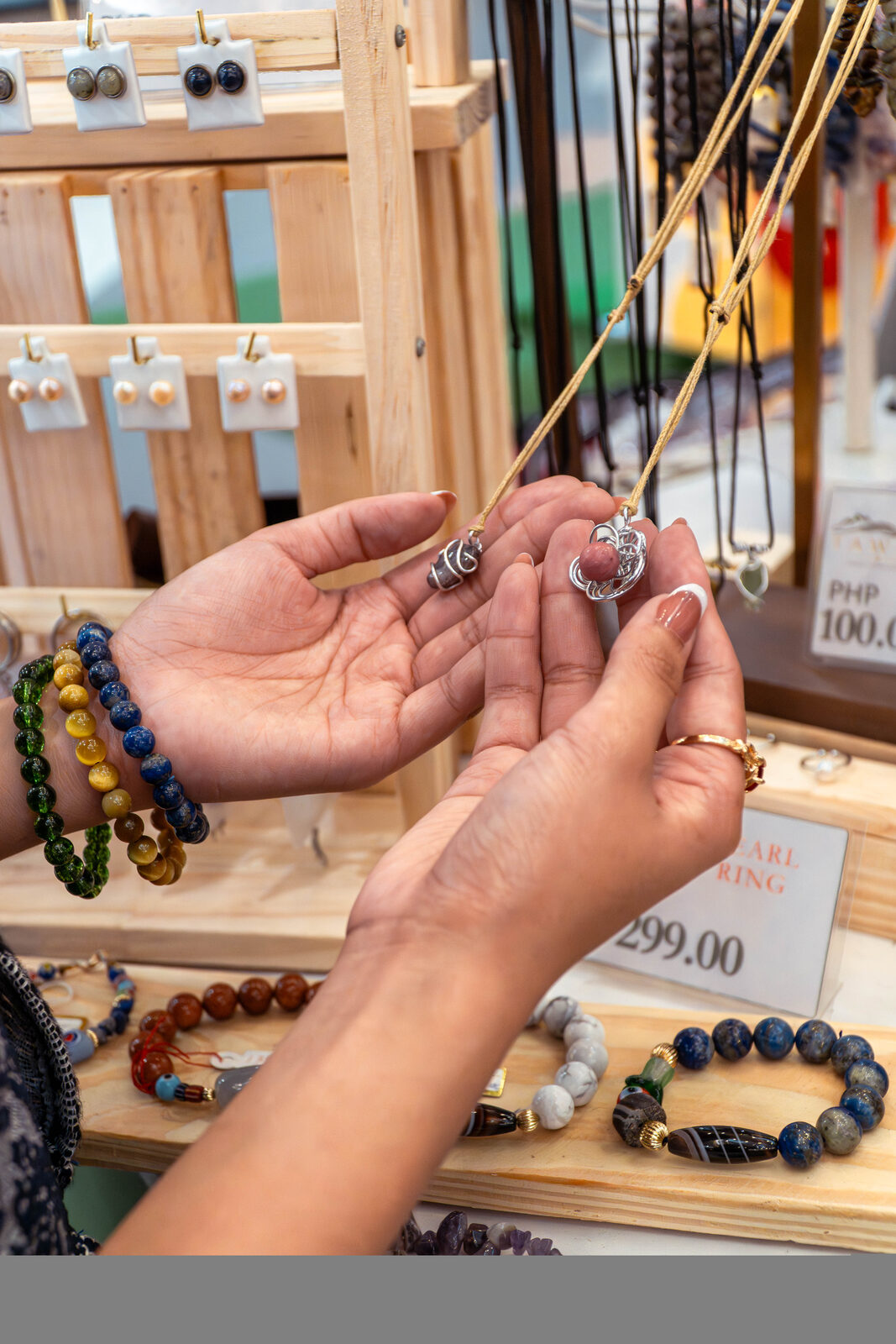 Hands comparing TÁWID™ stone pendants and bracelets at a market display.