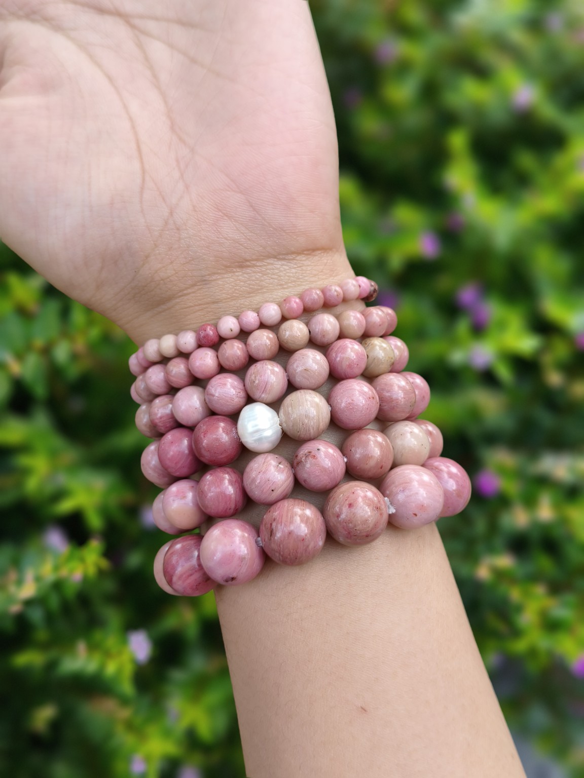 Pink rhodochrosite round-bead bracelets in graduated sizes, including a bracelet with a white pearl accent, worn on a wrist outdoors.